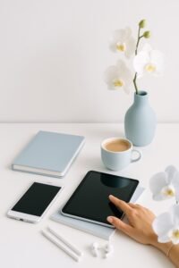 White orchids in a pale blue vase beside a blue notebook, coffee cup, phone and tablet on a white desk