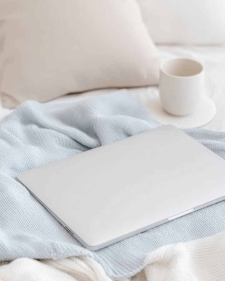 A white laptop and a cream cup resting on a soft blue knit blanket